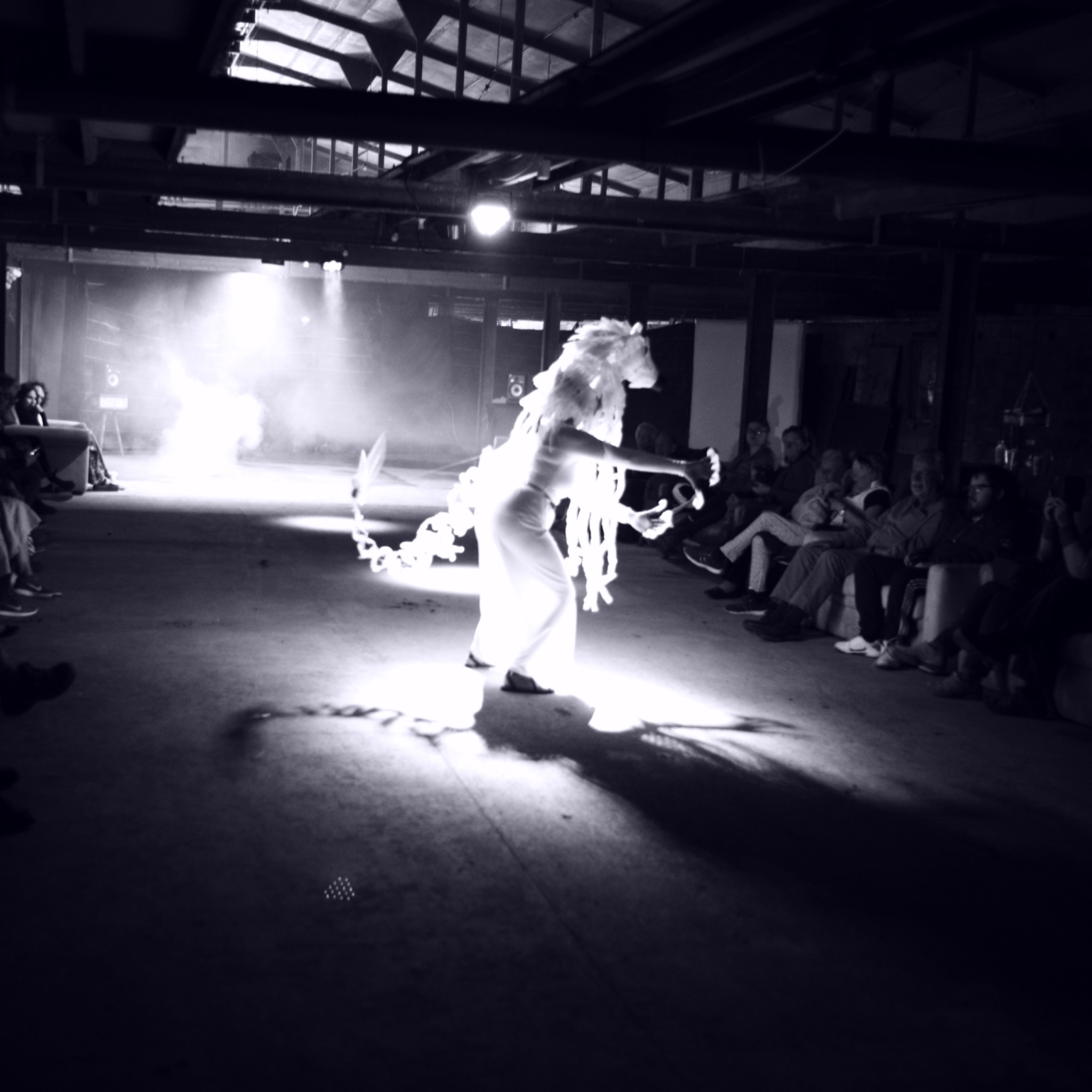 Sedna performance: artist in sculptural headpiece moving between seated audience in an industrial space, dramatic side-light.